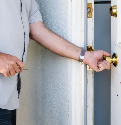 Close view of Jeff's hands at a white door: one on a brass doorknob, the other holding a small lock tool.