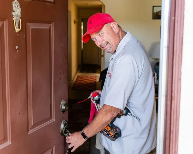 Jeff Slama at a brown front door, one hand at the lock and a cordless drill in the other, looking at the camera.